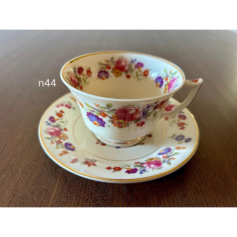 May include: A vintage teacup and saucer set. The white porcelain cup and saucer are adorned with a floral pattern in shades of red, purple, yellow, and blue. The rims are trimmed with gold. The text "n44" is visible on the side.