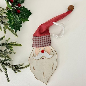 May include: A wooden Santa Claus Christmas ornament with a red hat and a white beard. The hat has a plaid pattern at the base. A small bell is attached to the end of the hat. The ornament is hanging by a white string.