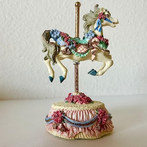 May include: A carousel horse figurine with a cream-colored body, adorned with blue and red floral accents. The horse has a flowing mane and tail, and is mounted on a gold-colored pole. The base is decorated with pink fabric and more floral details.
