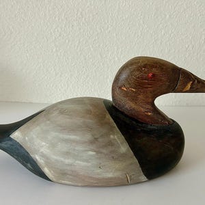 May include: A wooden duck decoy with a brown head and beak, a gray and white body, and a black tail. The duck has a red eye and is painted with visible wood grain.