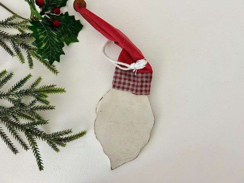 May include: A Santa Claus ornament with a red hat and a white beard. The hat is made of red fabric and has a checkered pattern at the base. A white string is attached to the hat. The beard is a light cream color.