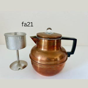 May include: Vintage copper coffee pot with a black handle and a glass knob on the lid. A separate metal filter basket is next to the pot. The text "fa21" is visible.
