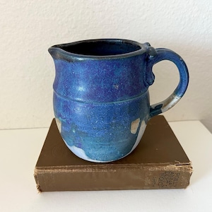 May include: A ceramic pitcher featuring a deep blue and turquoise glaze with visible white base areas. The pitcher has a rounded body, a curved handle, and a small pouring spout. It is resting on a brown hardcover book with visible text on the spine.