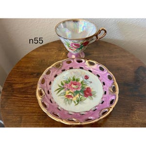 May include: A vintage teacup and saucer set. The iridescent teacup has a gold handle and is decorated with pink roses. The pink saucer has a gold trim and a floral center design. The text "n55" is visible on the left.