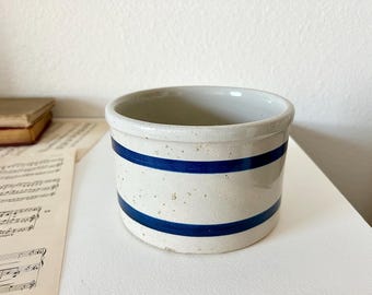 May include: A white ceramic crock with two blue bands around the top. The crock has a speckled texture and is perfect for storing small items.