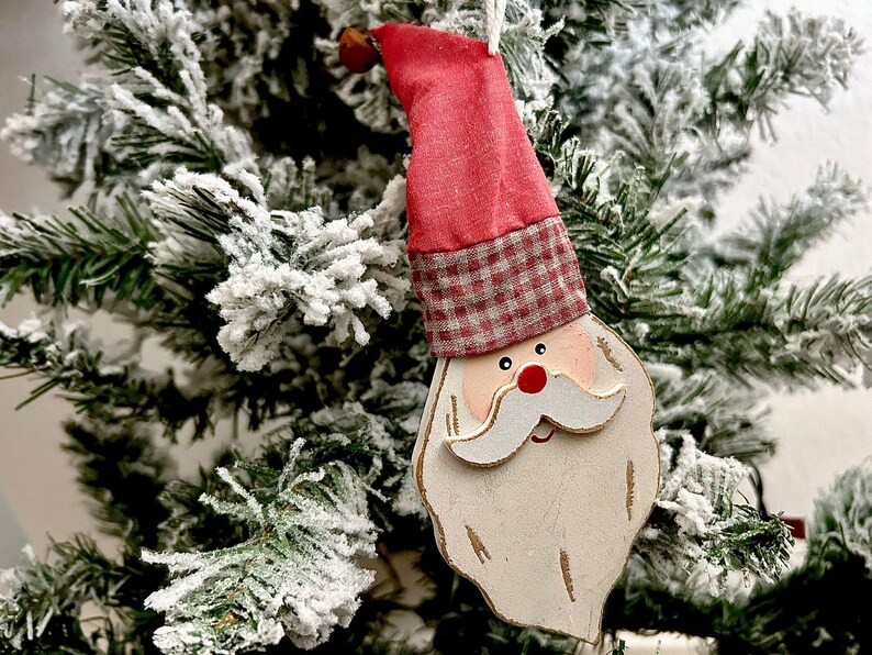 May include: A Santa Claus Christmas tree ornament. The ornament features a red hat with a plaid band, a white beard, and a red nose. The ornament is hanging in front of a snow-covered Christmas tree.