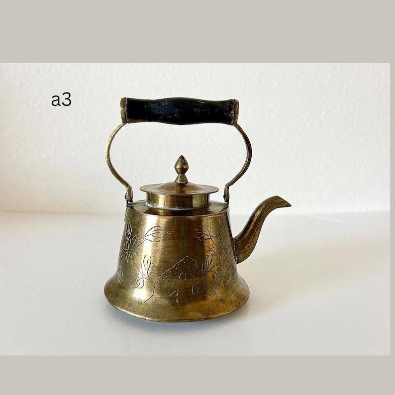 May include: Antique brass teapot with a black wooden handle. The teapot has a curved spout and a lid with a small finial. The body of the teapot is decorated with an etched floral design. The text "a3" is visible in the image.