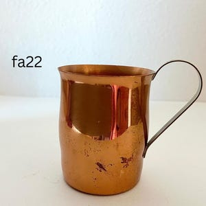 May include: A copper mug with a curved handle. The mug has a cylindrical shape with a slightly flared rim. The surface of the mug has a warm, reddish-brown color with some darker spots, and the handle is a darker metal.