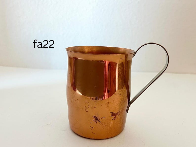 May include: A copper mug with a curved handle. The mug has a cylindrical shape with a slightly flared rim. The surface of the mug has a warm, reddish-brown color with some darker spots, and the handle is a darker metal.