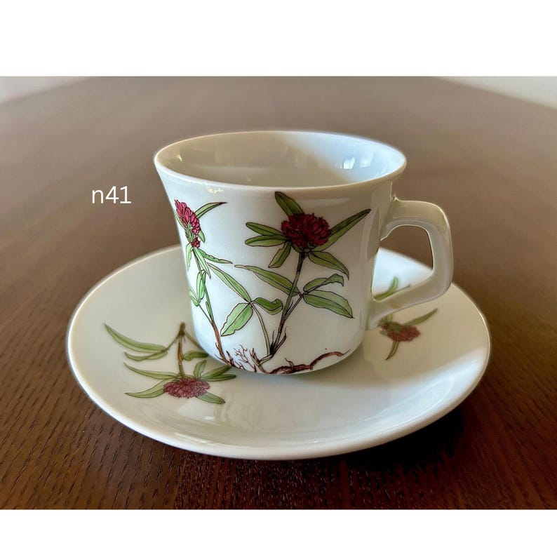 May include: White teacup and saucer set with a floral design featuring red flowers and green leaves. The cup has a handle and sits on a matching saucer. The set is on a wooden surface.