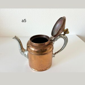 May include: Copper teapot with a silver handle and spout, and a hinged lid. The teapot has a decorative design around the top and base. The lid is open, revealing the interior. The teapot is on a white surface.