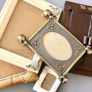 May include: A collection of decorative picture frames in various styles. Includes a square cream-colored frame with floral accents, a rectangular woven frame, and a dark brown wooden frame. The central frame is ornate with an oval opening.