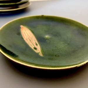 May include: A set of green ceramic plates with a gold rim. Each plate features a stylized, light orange leaf design. The plates have a glossy finish and are suitable for serving appetizers or small dishes.