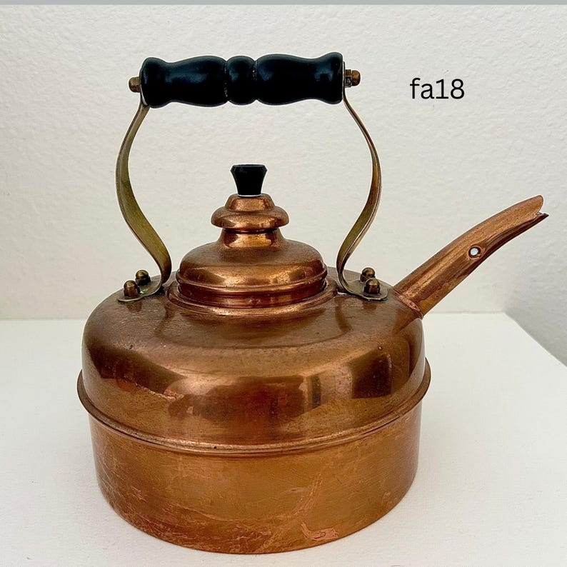 May include: A vintage copper tea kettle with a black wooden handle and spout. The kettle has brass accents and a small black knob on the lid. The text "fa18" is visible on the side of the kettle.