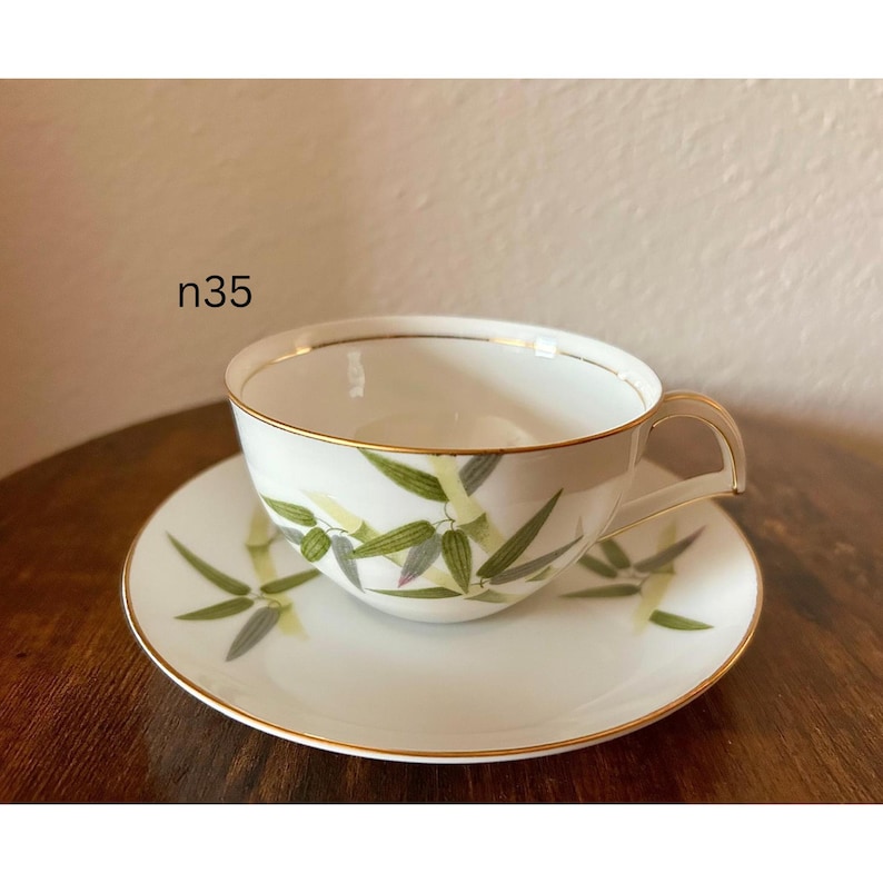 May include: White teacup and saucer set with a gold rim and bamboo leaf design. The teacup has a curved handle and sits on a matching saucer. The text "n35" is visible on the image.