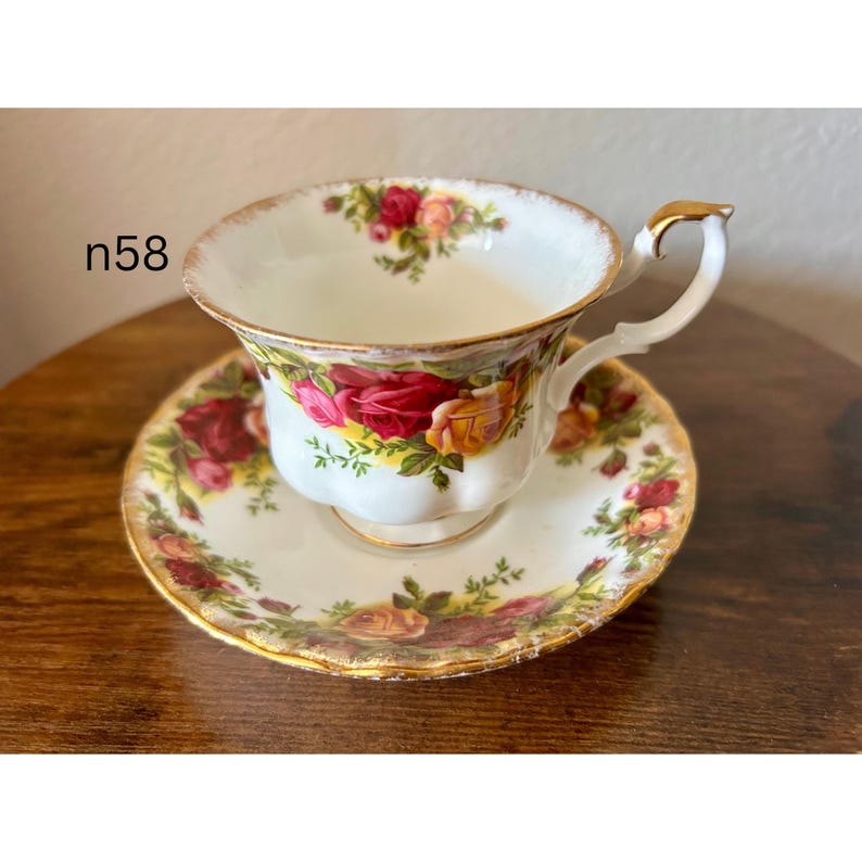 May include: A white teacup and saucer set with a floral rose pattern in red, yellow, and green. The cup has a gold handle and trim. The saucer is round with a matching floral design. The text "n58" is visible on the left side.