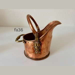 May include: A small, antique copper bucket with a handle and spout. The bucket has a brass handle and a decorative brass emblem on the side. The text "fa36" is visible on the side of the bucket.