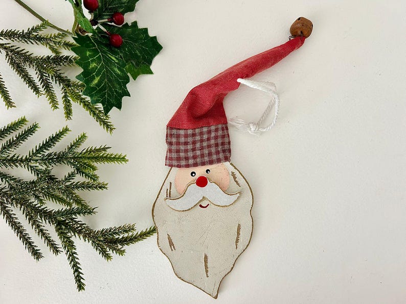 May include: A wooden Santa Claus Christmas ornament with a red hat and a white beard. The hat has a plaid pattern at the base. A small bell is attached to the end of the hat. The ornament is hanging by a white string.