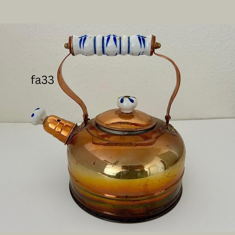 May include: A copper-colored tea kettle with a rounded body and a curved handle. The handle is copper with a white and blue ceramic grip. The spout and lid also have white and blue ceramic accents. The kettle is on a white surface.