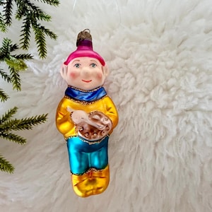 May include: A vintage Christmas ornament shaped like a smiling artist. The ornament features a pink hat, a yellow jacket, blue pants, and holds a brown palette. The ornament is on a white, fluffy surface.