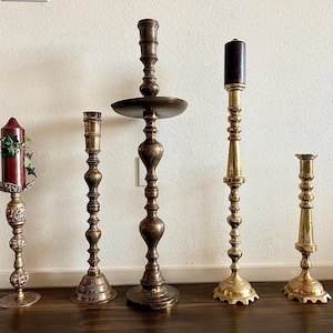 May include: Five ornate brass candle holders with varying heights and designs. The candle holders are on a light brown wooden surface.