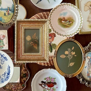 May include: A collection of decorative items including a rectangular gold frame with pressed leaves, an oval gold frame with a pressed plant, two white plates with bird illustrations, a blue and white plate, and various framed wall art pieces.