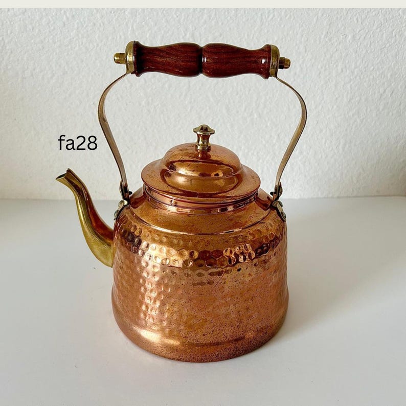 May include: A copper kettle with a hammered finish, featuring a wooden handle and brass accents. The spout and handle are brass. The kettle has a rounded body and a lid with a small knob. The text "fa28" is visible on the side.