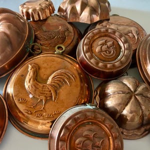 May include: A collection of various copper baking molds featuring embossed designs including a rooster, a sailing ship, and floral patterns. These metallic kitchen tools have rounded shapes and hanging loops for display or storage purposes.