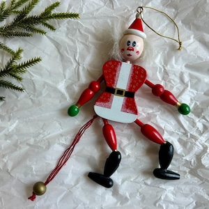 May include: A wooden Santa Claus jumping jack ornament featuring a red and white suit, black belt, and black boots. The figure has a painted face, white hair, and green-tipped hands. A red string with a gold bead hangs from the bottom for movement.