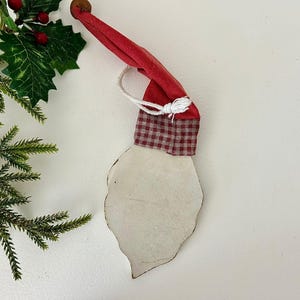May include: A Santa Claus ornament with a red hat and a white beard. The hat is made of red fabric and has a checkered pattern at the base. A white string is attached to the hat. The beard is a light cream color.
