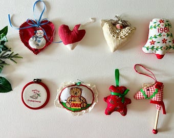 May include: A collection of handmade Christmas ornaments. The ornaments include a snowman heart, a bell with "1981", a bird in a nest, a teddy bear playing drums, and a red ornament shaped like a bear. Each ornament has a loop for hanging.