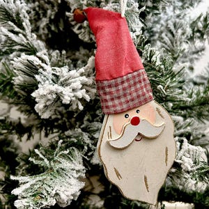 May include: A Santa Claus Christmas tree ornament. The ornament features a red hat with a plaid band, a white beard, and a red nose. The ornament is hanging on a snow-covered Christmas tree.