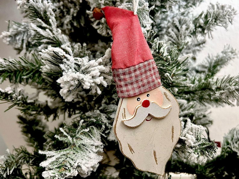 May include: A Santa Claus Christmas tree ornament. The ornament features a red hat with a plaid band, a white beard, and a red nose. The ornament is hanging on a snow-covered Christmas tree.
