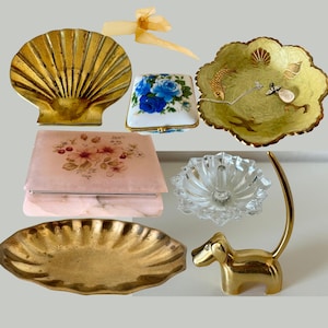 May include: A collection of decorative trinket dishes and trays.  A gold shell-shaped dish, a green and gold dish with a fish design, a pink marble box with floral designs, a clear glass ring holder, a gold oval dish, and a gold dachshund ring holder.