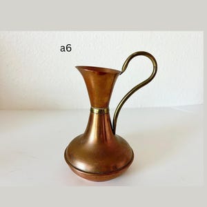 May include: A copper-toned pitcher with a brass handle and rim. The pitcher has a bulbous base and a flared top. The handle curves gracefully from the top to the side. The text "a6" is visible in the upper left corner.