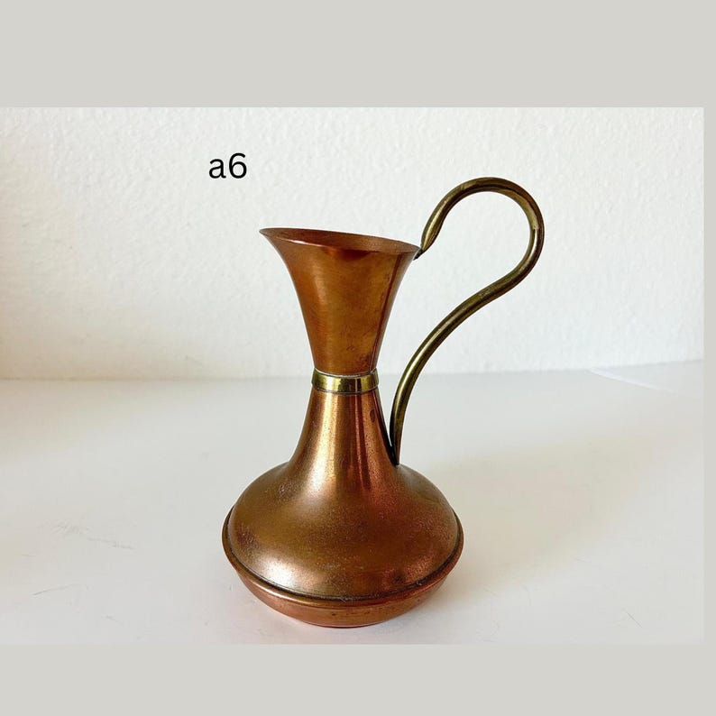 May include: A copper-toned pitcher with a brass handle and rim. The pitcher has a bulbous base and a flared top. The handle curves gracefully from the top to the side. The text "a6" is visible in the upper left corner.