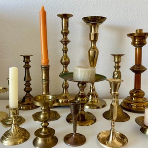 May include: A collection of brass and gold-toned candlesticks of various sizes and designs. Some hold lit or unlit candles in shades of orange and white. The candlesticks are arranged on a white surface.