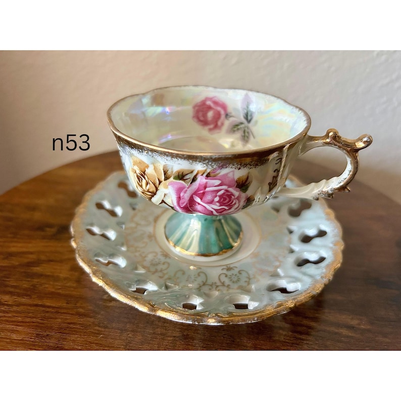 May include: Antique teacup and saucer set. The teacup features a floral design with pink roses and gold trim. The saucer has a delicate, openwork design with gold edging. The teacup is supported by a pedestal base with a turquoise accent. The text "n53" is visible.
