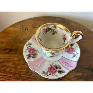 May include: A vintage teacup and saucer set. The cup and saucer are white with pink and gold accents. The saucer has a pink and white striped pattern with pink roses. The cup has a gold rim and a handle.