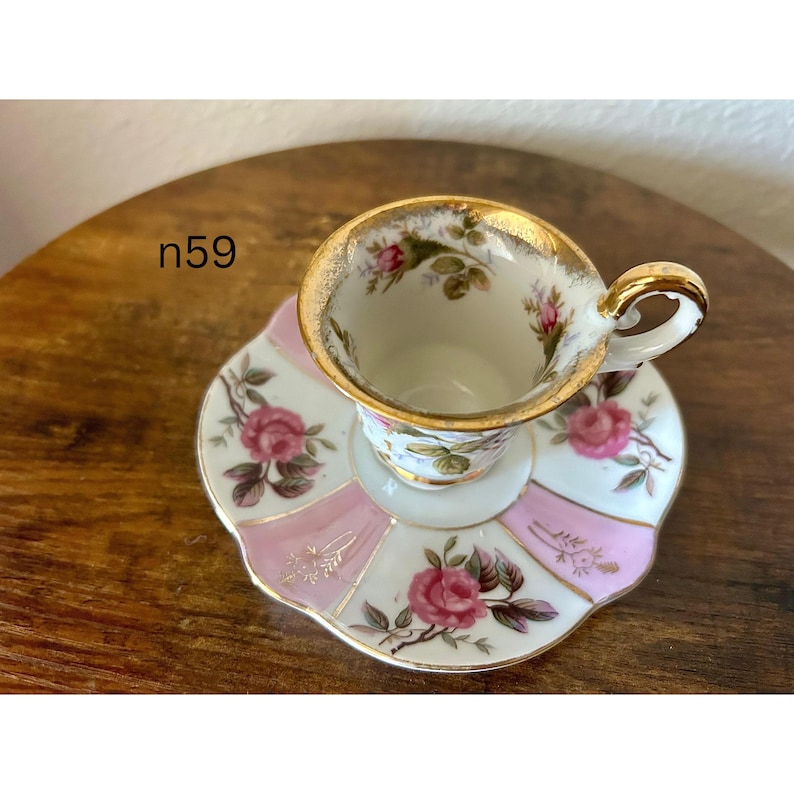 May include: A vintage teacup and saucer set. The cup and saucer are white with pink and gold accents. The saucer has a pink and white striped pattern with pink roses. The cup has a gold rim and a handle.