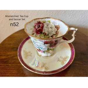 May include: A mismatched tea cup and saucer set. The cup and saucer are white with a gold trim and a floral design featuring red, white, and blue roses. The saucer has a pink border and gold accents. Text on the image reads "Mismatched Tea Cup and Saucer Set" and "n52".