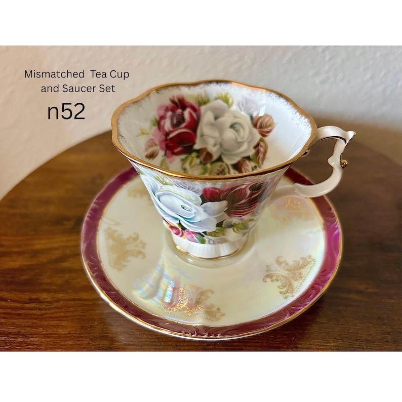 May include: A mismatched tea cup and saucer set. The cup and saucer are white with a gold trim and a floral design featuring red, white, and blue roses. The saucer has a pink border and gold accents. Text on the image reads "Mismatched Tea Cup and Saucer Set" and "n52".