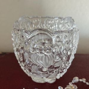 May include: A clear glass candle holder with a raised design of a Santa Claus figure. The holder has a scalloped rim and is perfect for adding a festive touch to your home decor.