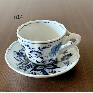 May include: White teacup and saucer set with a blue floral pattern. The cup has a unique handle and a scalloped rim. The saucer also features the same blue floral design. The set is on a wooden surface.