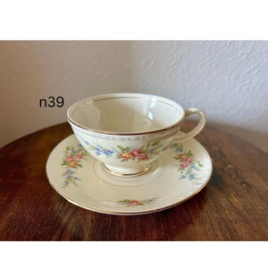 May include: Cream-colored teacup and saucer set with floral designs in pink, yellow, and blue. The cup and saucer have gold trim. The text "n39" is in the upper left corner.