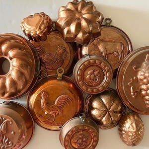 May include: A collection of antique copper kitchen molds in various shapes, including a heart, a flower, a rooster, and a bundt cake. The molds are arranged on a white surface, showcasing their intricate designs and warm, metallic color.