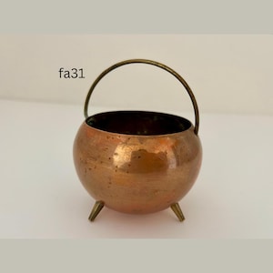 May include: A small, round copper cauldron with a brass handle and three small legs. The cauldron has a shiny, reflective surface and is likely used for decorative purposes. The text "fa31" is visible on the side.