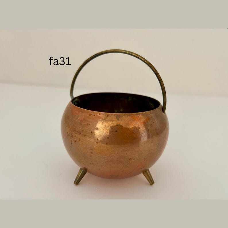 May include: A small, round copper cauldron with a brass handle and three small legs. The cauldron has a shiny, reflective surface and is likely used for decorative purposes. The text "fa31" is visible on the side.