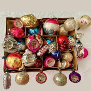 May include: A collection of assorted vintage glass Christmas ornaments in a cardboard box. The ornaments feature various shapes, including spheres, teardrops, and indented designs, with metallic, glossy, and matte finishes in red, gold, blue, and silver.