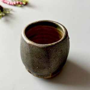 May include: A handmade, small, ceramic cup with a dark, textured exterior. The interior is a deep brown, and the rim has a subtle green hue. The cup has a rustic, organic aesthetic, perfect for beverages or decorative use.